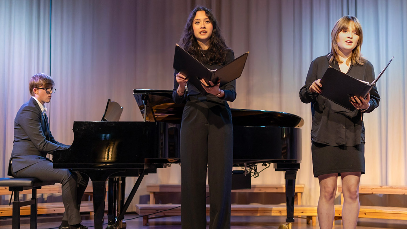 Två kvinnor sjunger och en man spelar piano på scen.