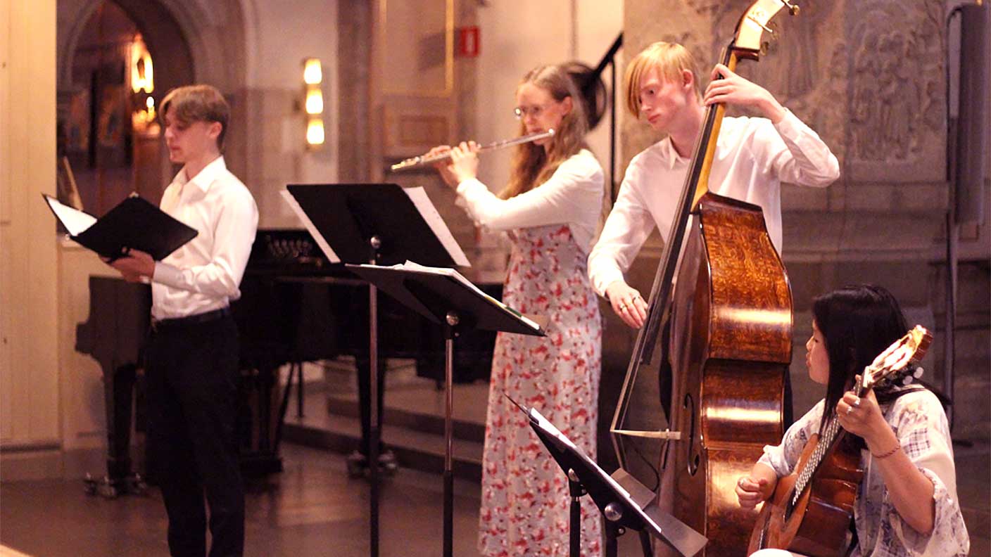 Personer framför musik på gitarr, kontrabas, tvärflöjt och sång i kyrkomiljö.