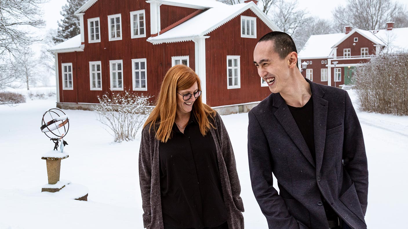 Andreas och rektor Lisa står bredvid varandra i ett snöigt Kävesta-landskap med ett rött hus med vita knutar i bakgrunden. De är båda klädda i grått och svart. Andreas i t-shirt och kavaj, Lisa i skjorta och lång kofta. De ler och skrattar.