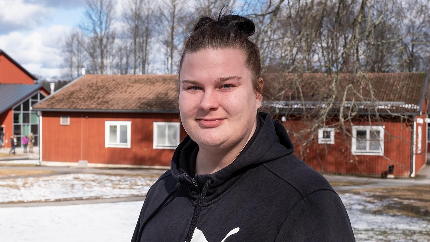 Emma står på en snötäckt gräsmatta och tittar in i kameran. I bakgrunden syns två röda hus.