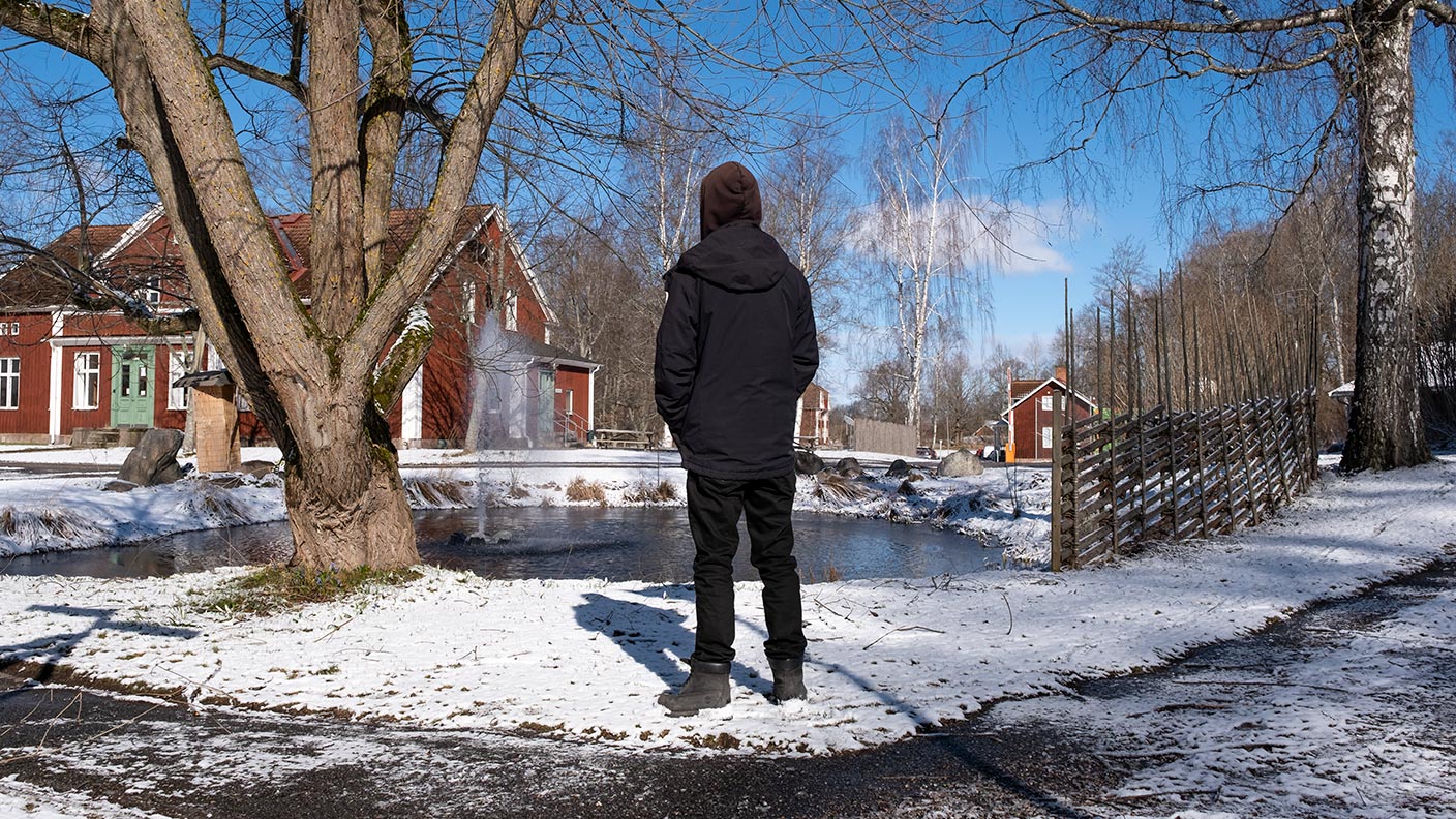 Gabriel står mitt i bilden med ryggen vänd mot kameran. Gabriel tittar ut över en damm och några röda byggnader. Det är snö på marken.