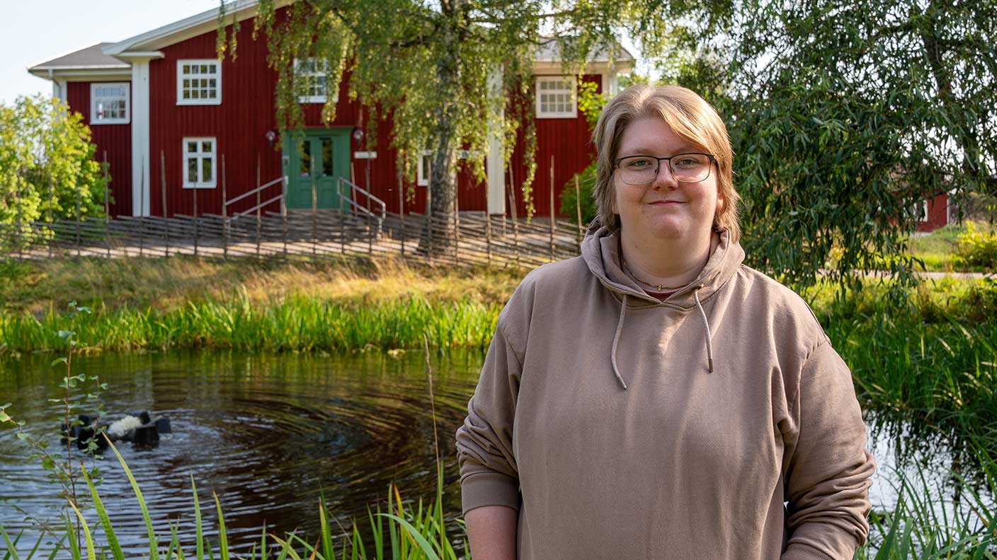 Kvinna i halvprofil med kort, rakt, ljusbrunt hår, glasögon och brun huvtröja. Hon ler mot kameran. I bakgrunden en damm, lummig grönska och ett rött trähus.