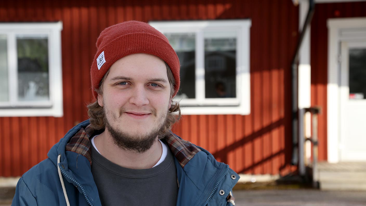 Oskar Eksell står framför en av Kävestas röda träbyggnader. Han bär en öppen jacka och röd mössa. På marken ligger snö.