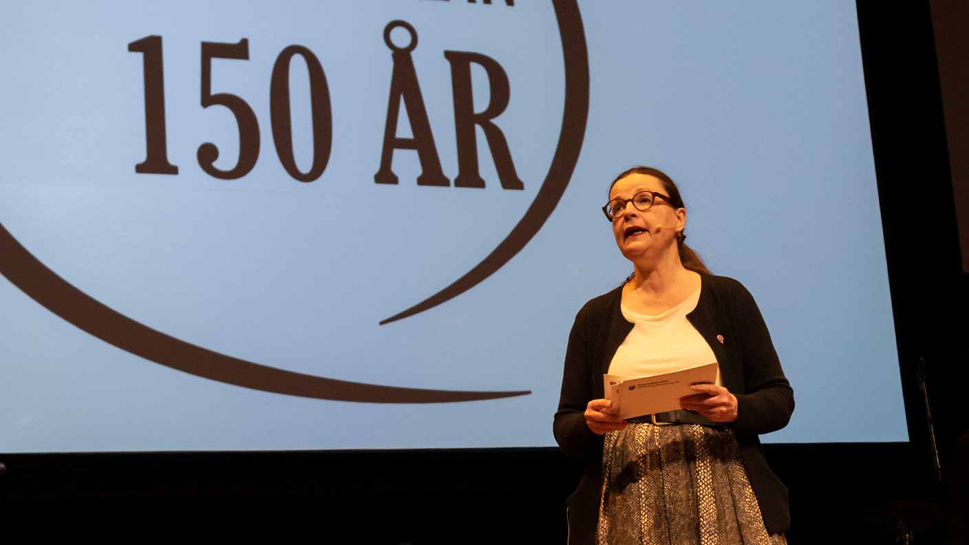 Anna Ekström, klädd i vit blus, svart uppknäppt kofta och brun mönstrad långkjol, framför en stor skärm där man ser texten "150 år". Anna håller i ett manuskort och ser ut att prata till en publik. 