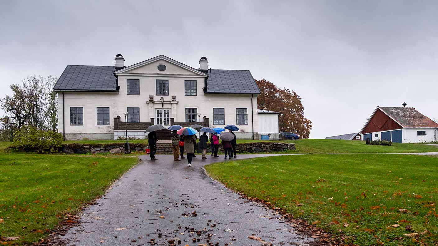 En lite grupp personer står med paraplyer på uppfarten till en stor vit mangårdsbyggnad - Vrana gård.