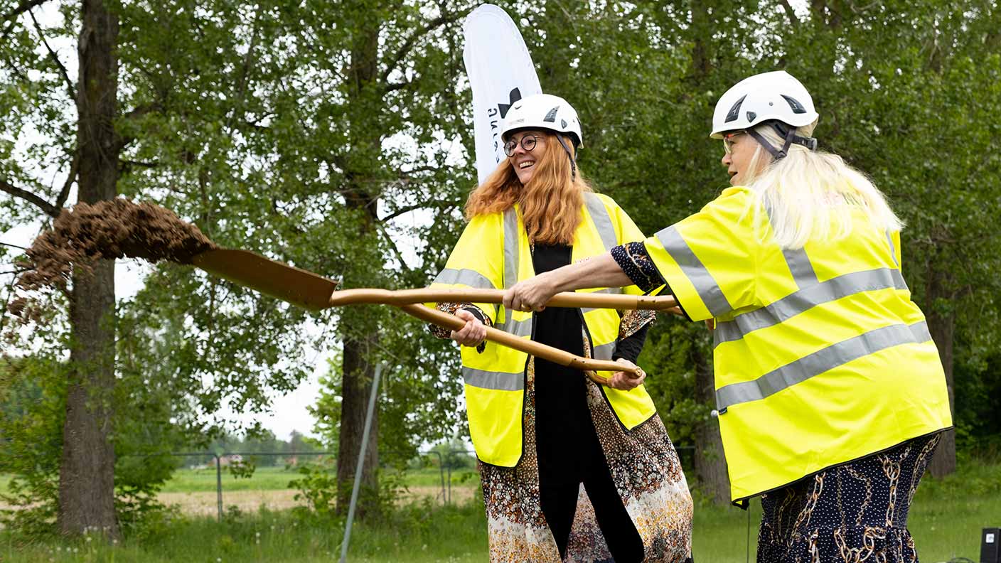 Två personer med gula varselvästar och skyddshjälmar använder en spade med två handtag för att skyffla sand.