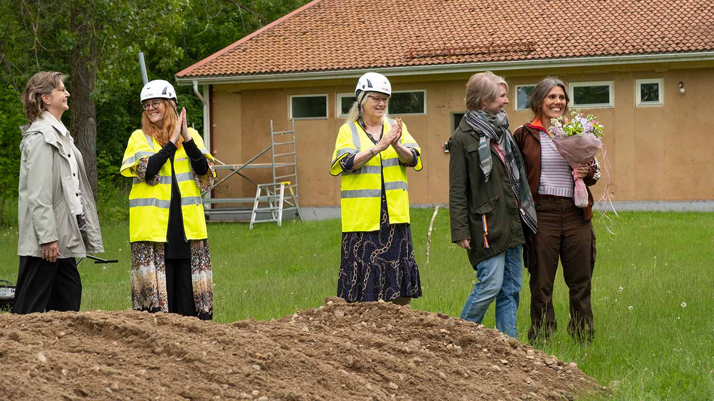 Bakom en hög sand står fem personer, varav två i gula vaserlvästar och skyddshjälmar. Två av personerna är mer ledigt klädda och håller om varandra med en arm. En av dem håller i en blombukett. I bakgrunden syns ett hus som är i färd att få ny fasad.