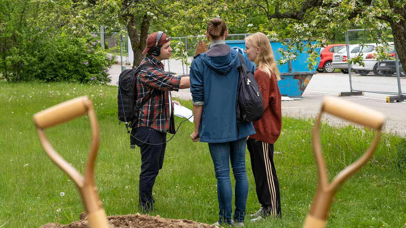 Två personer intervjuas av en radioreporter som bär en ryggsäck kopplad till ett headset och en handmikrofon.