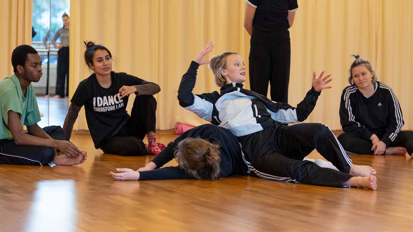 En grupp dansare i en danssal. Tre sitter på golvet och tittar på två andra, varav en ligger utsträckt med den andra personen sittande och gestikulerande på sig. Alla bär träningskläder.