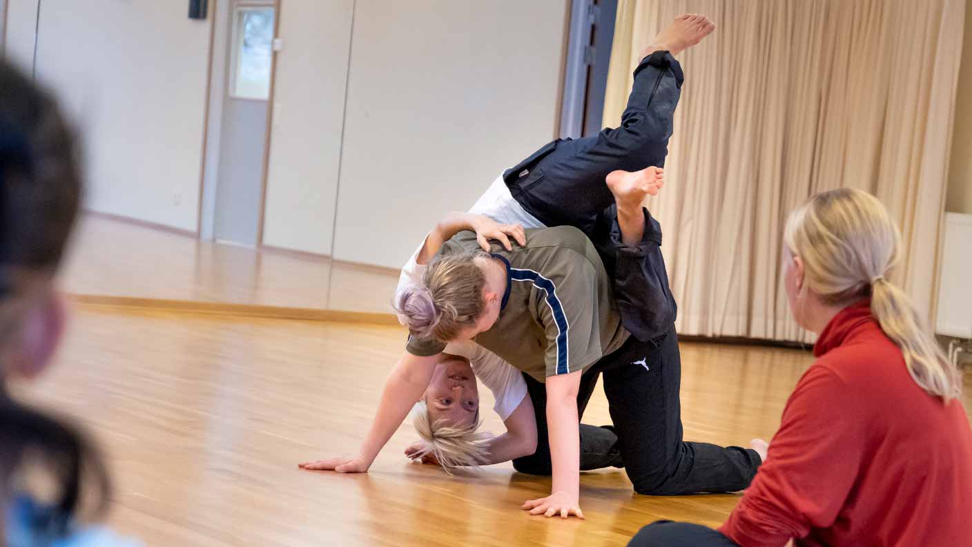 Två personer är inbegripna i en akrobatisk manöver. Den ena står stadigt på alla fyra. Den andra har klättrat upp på den förstes rygg och hänger ner vid sidan av ryggen, samtidigt som den tittar på några åskådare under magen och mellan armarna på den första personen.