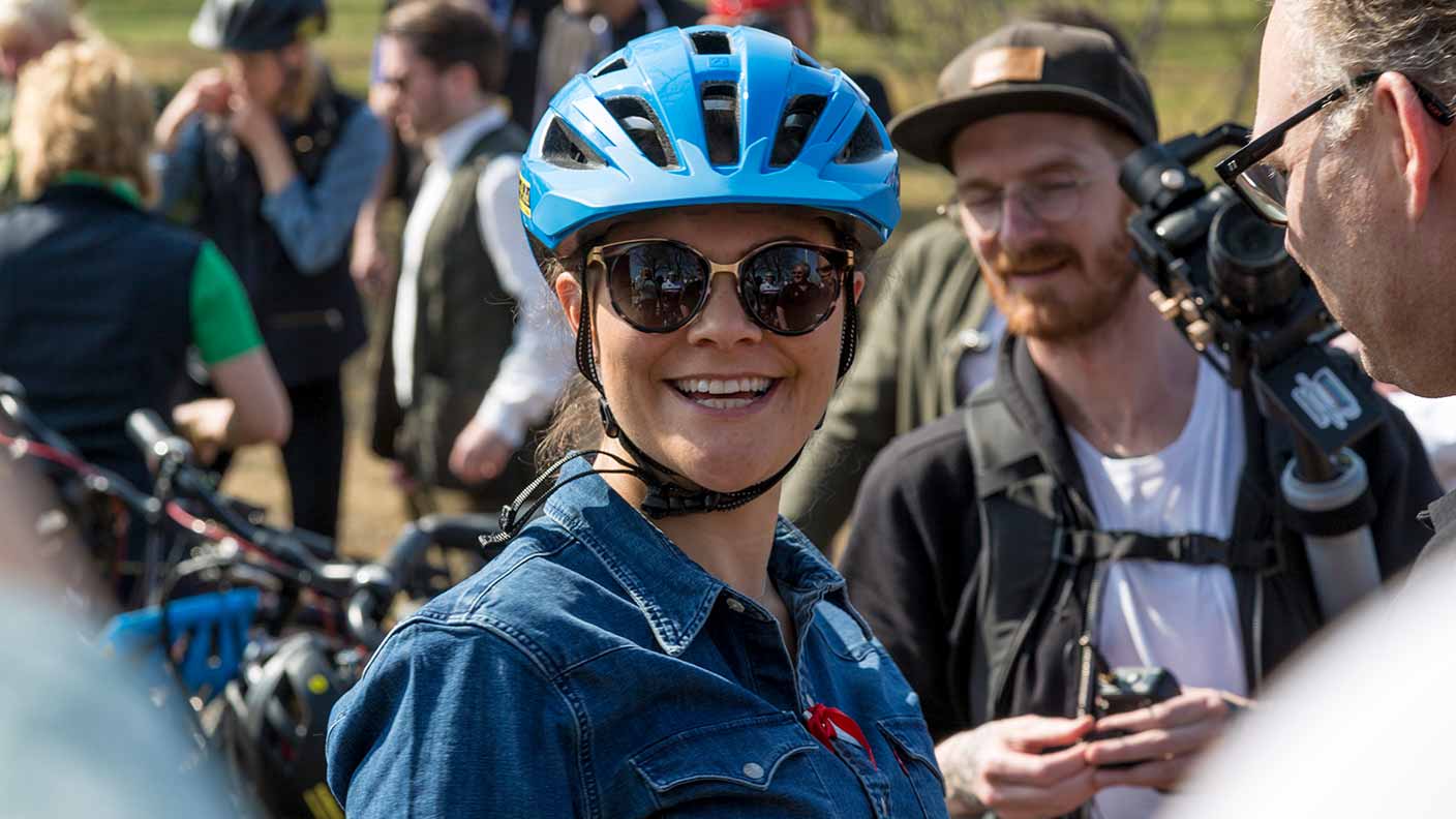 Kronprinsessan iförd blå cykelhjälm, mörka solglasögon och blå jeanströja. Hon ler och tittar rakt mot kameran. Runt omkring syns personer som fotograferar, tittar på eller väntar med sina cyklar.