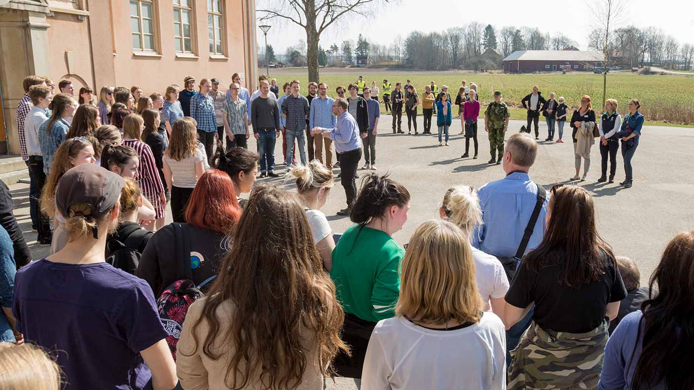 Kronprinsessan står i en liten grupp människor och lyssnar till en kör som står framför Kävestas stora skolhus, en orange stenbyggnad. I bildens förgrund syns ett myller av åskådare.