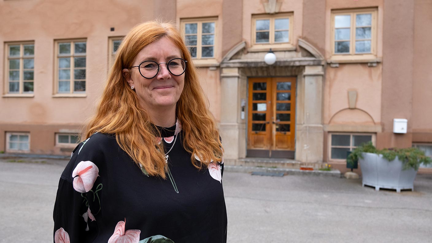 Lisa Rosander, klädd i svart klänning med stora blommönster, står framför stora skolhuset på Kävesta folkhögskola