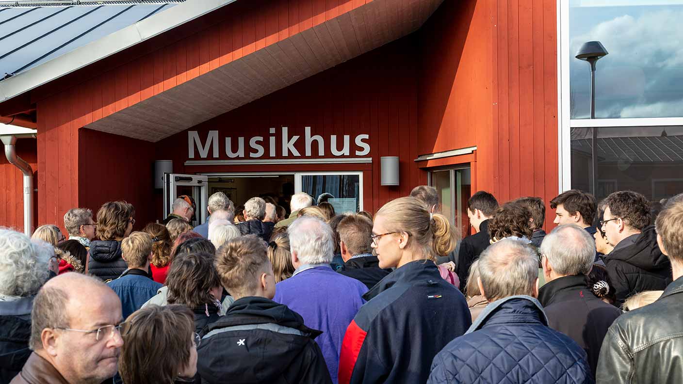 En stor folkmassa rör sig mot dörrarna till ett rött hus. Ovanför dörren står det "Musikhus" med stora vita bokstäver.