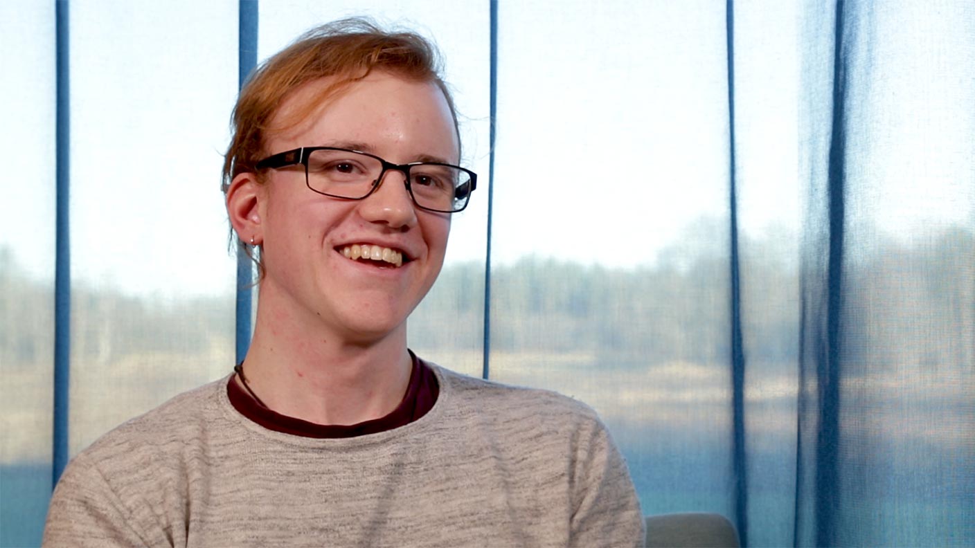 Justus sitter på en stol framför ett stort panoramafönster som täcks av en delvis genomskinlig blå gardin. Justus ler och tittar snett in i kameran. Han har röd-orange hår, svarta glasögon och bär en grå långärmad tröja.