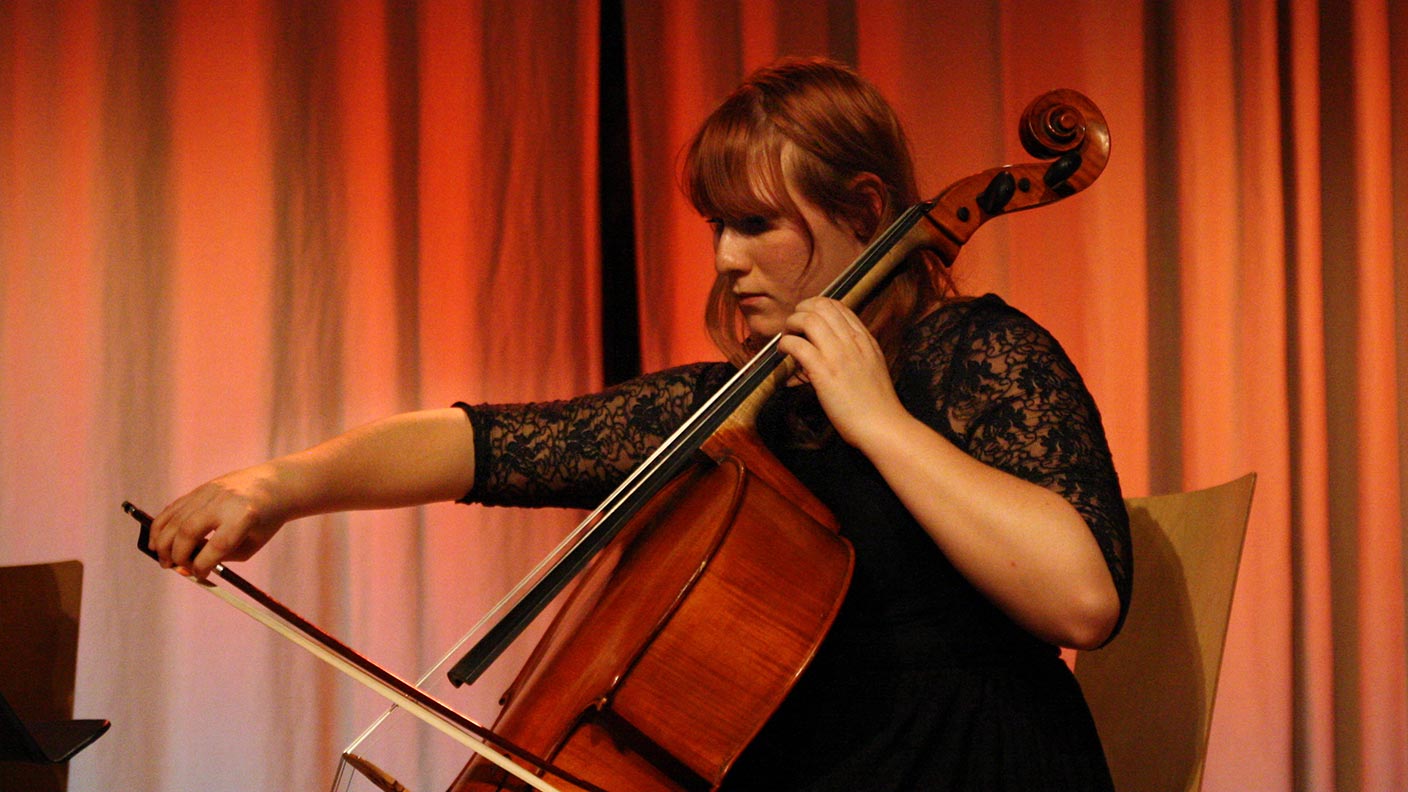 Sofie spelar cello på en scen. Hon sitter på en stol och är klädd i en svart klänning.