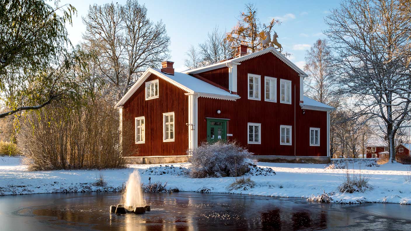 Rött trähus med vita knutar i vinterlandskap. Damm med fontän i förgrunden.