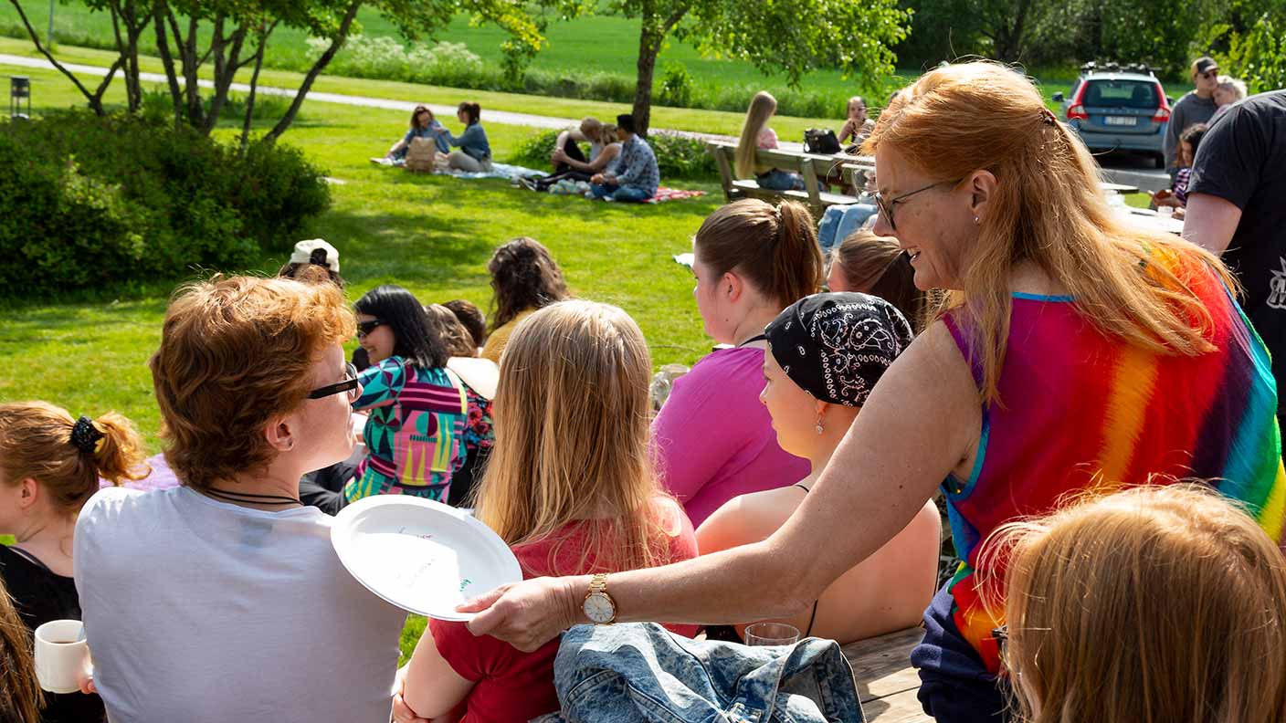 Skolans rektor Lisa, i regnbågsfärgad klänning, pratar med en deltagare i vit t-shirt. Det är en solig och grön försommardag och runt paret sitter och rör sig många andra deltagare och personal.