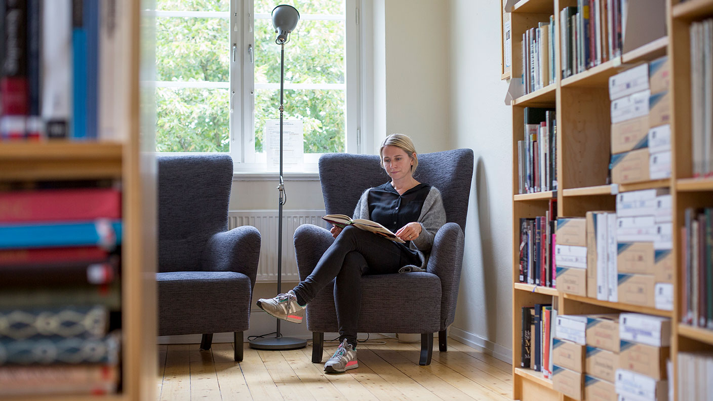 En person sitter i en stor fåtölj framför ett fönster och läser en bok. I förgrunden syns bokhyllor på både vänster och höger sida.