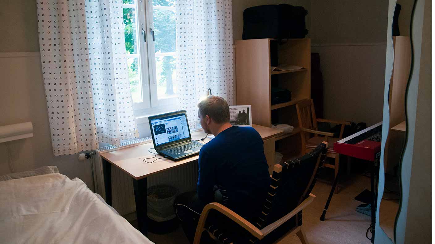 En person sitter i framför ett bord med en laptop i sitt rum. Bordet står framför ett högt fönster. I rummet finns också en säng, hyllor och ett elpiano.