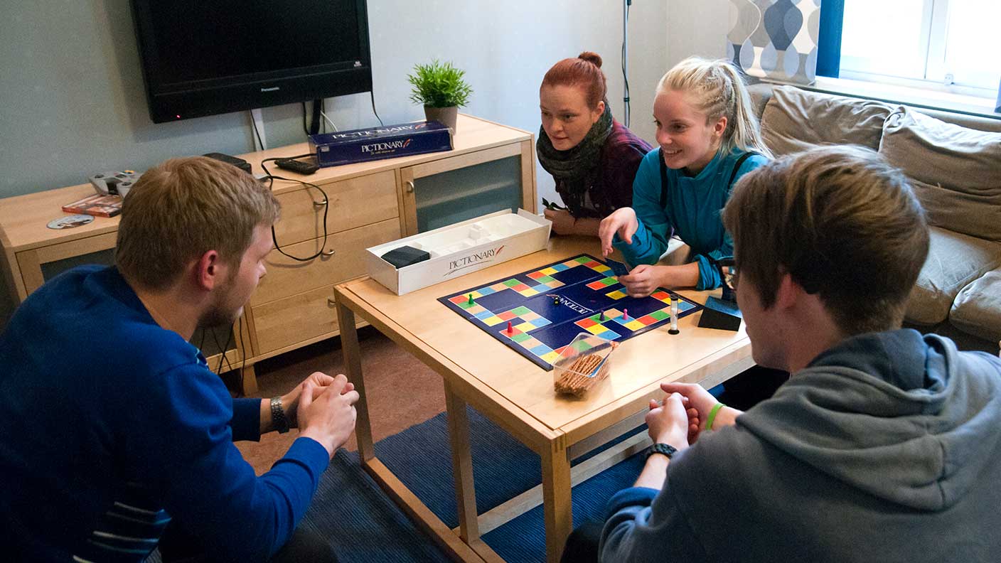 Fyra personer sitter i ett vardagsrum med soffa, fåtöljer, bord och en TV. De är samlade kring bordet och spelar ett brädspel.