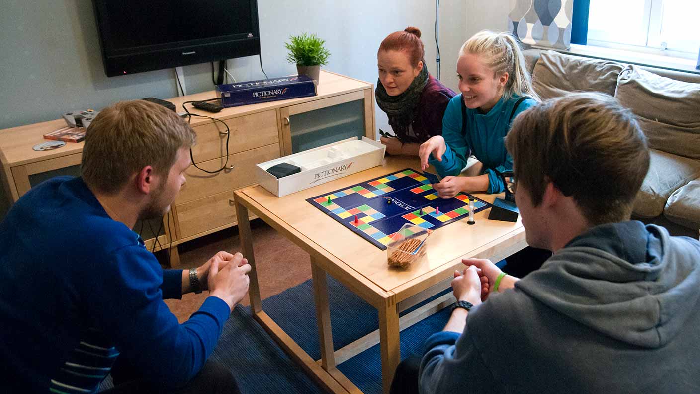 Fyra personer spelar spel vid ett litet kvadratiskt bord. I bakgrunden syns en vägghängd TV.