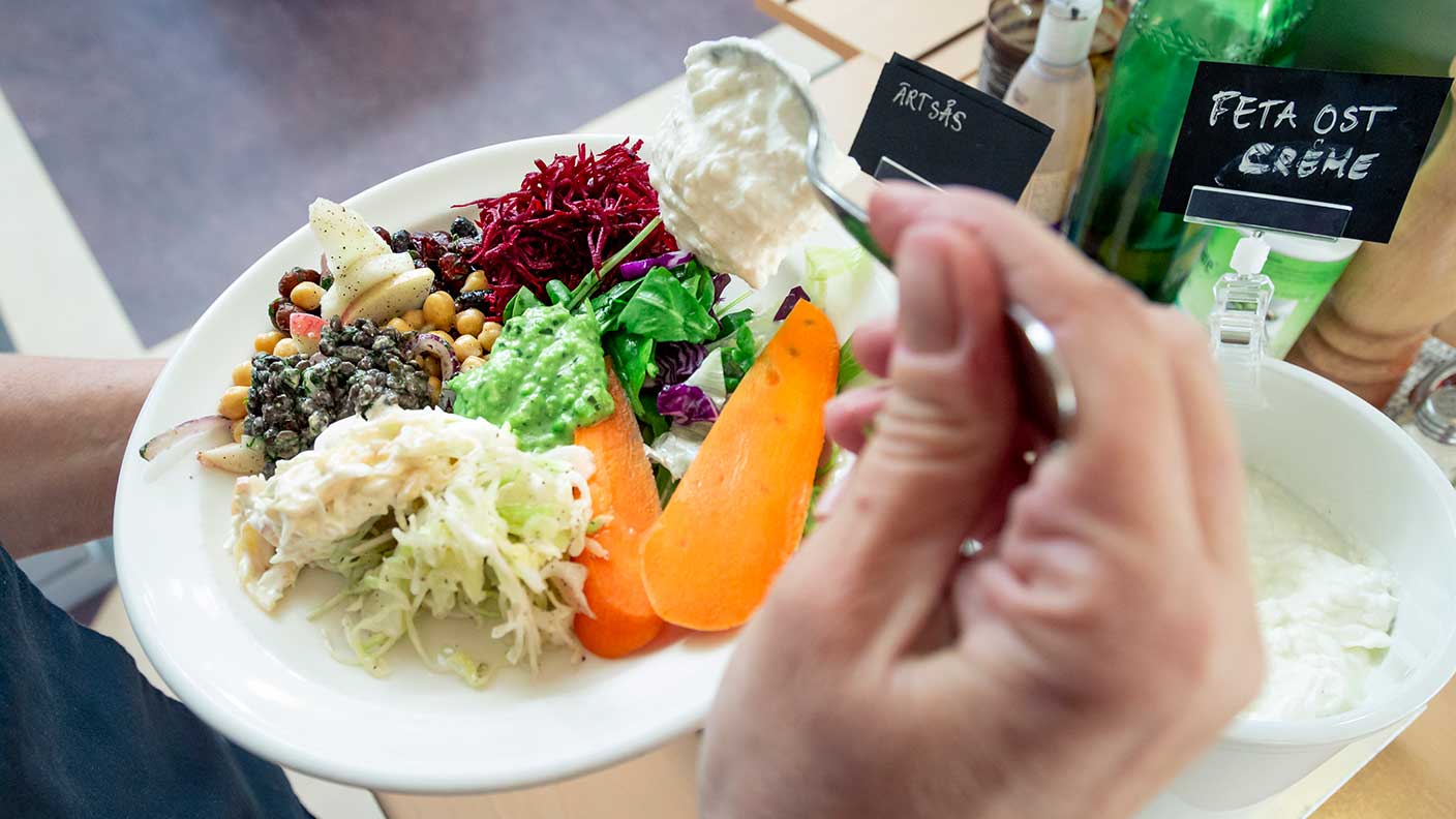 En hand lägger en klick fetaostås på en tallrik med sallad - morötter, kålsallad, rödbetor med mera.