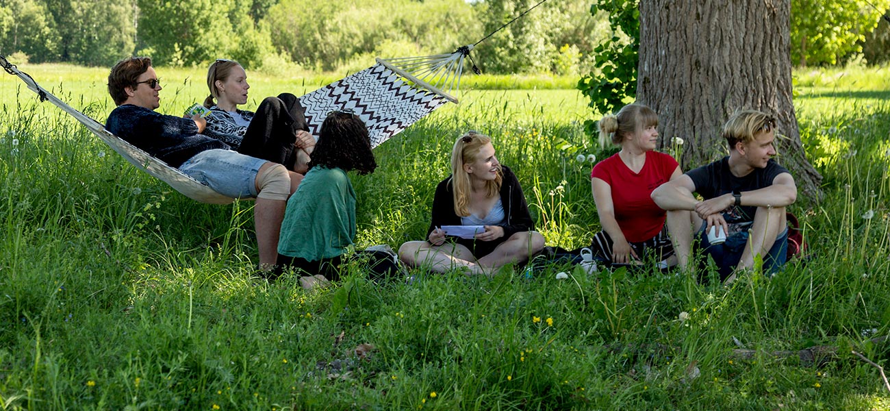 Sex personer ha samlats i en grupp ute i grönskande sommarväder. Två sitter i en hängmatta och övriga i gräset. Några tittar på något som pågår utanför bild, några talar med varandra.