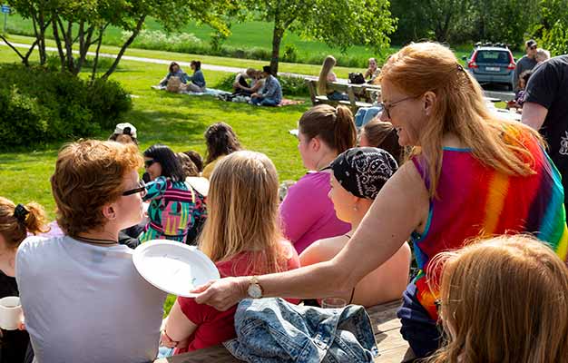 Skolans rektor Lisa, i regnbågsfärgad klänning, pratar med en deltagare i vit t-shirt. Det är en solig och grön försommardag och runt paret sitter och rör sig många andra deltagare och personal.