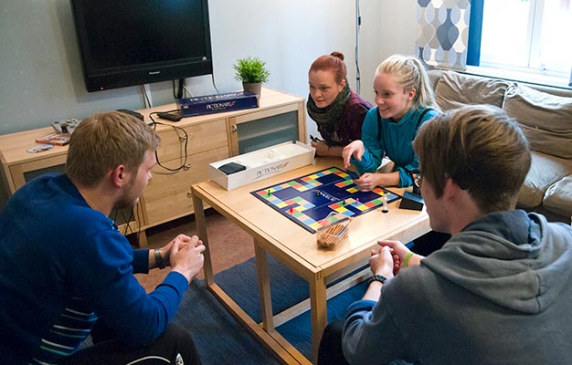 Fyra personer sitter i ett vardagsrum med soffa, fåtöljer, bord och en TV. De är samlade kring bordet och spelar ett brädspel.