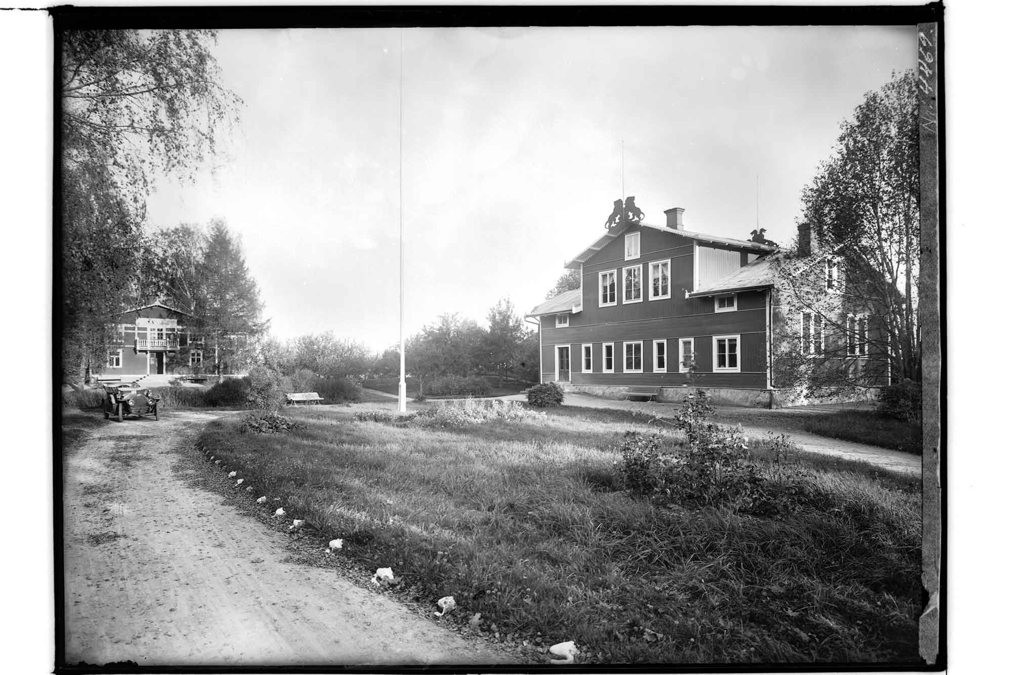 En grusväg leder in mot två hus. Det som ligger längst bort skyms av träd. Till höger i bild syns tydligt ett större tvåvåningshus med lejonornament på taket.