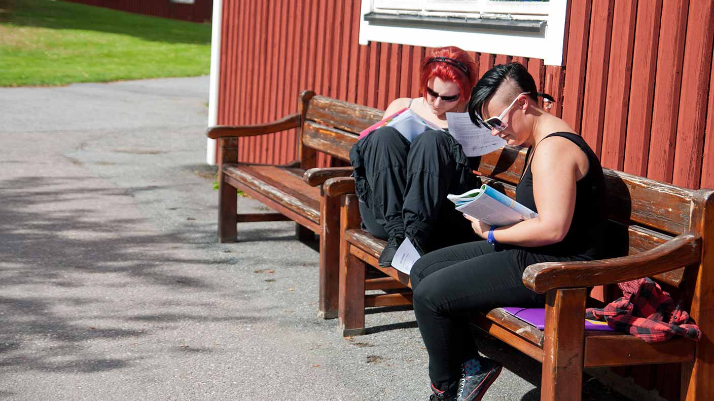 Två deltagare sitter avslappnat på en bänk utanför en röd byggnad och läser var sin bok.