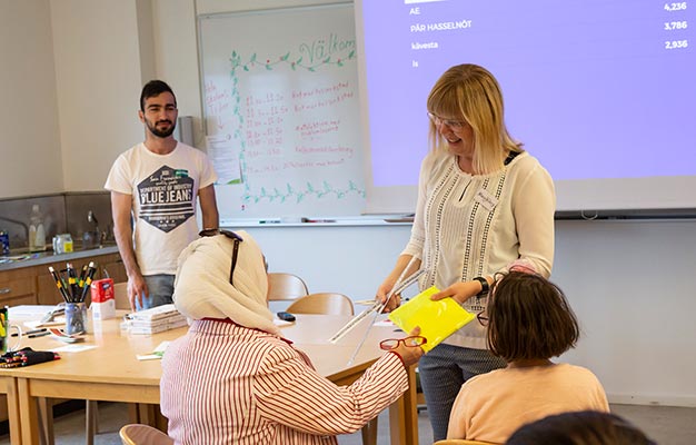 En lärare i ljust axellångt hår och vit tröja räcker över en gul reflexväst till den deltagare som bär vit sjal och en röd- och vitrandig kavaj.