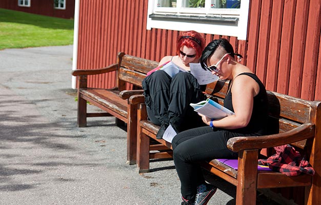 Två deltagare sitter avslappnat på en bänk utanför en röd byggnad och läser var sin bok.