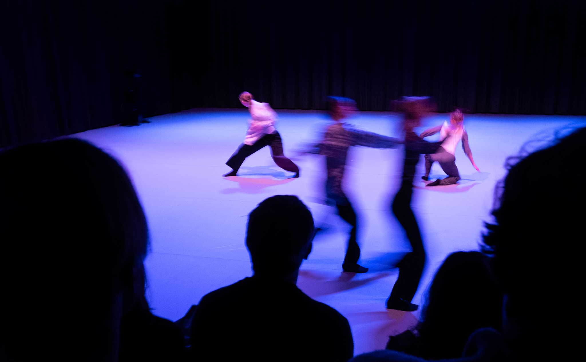 En dansföreställning sedd över axlarna på flera i publiken. På scengolvet rör sig en grupp dansare så snabbt att rörelseoskärpan gör dem suddig.