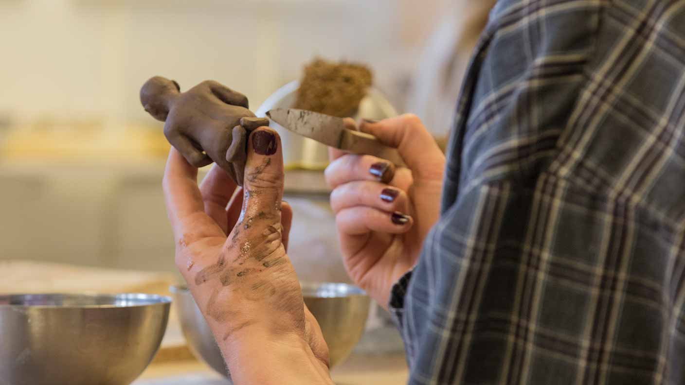 Närbild på en persons händer, som arbetar med en keramikskulptur i brun lera.