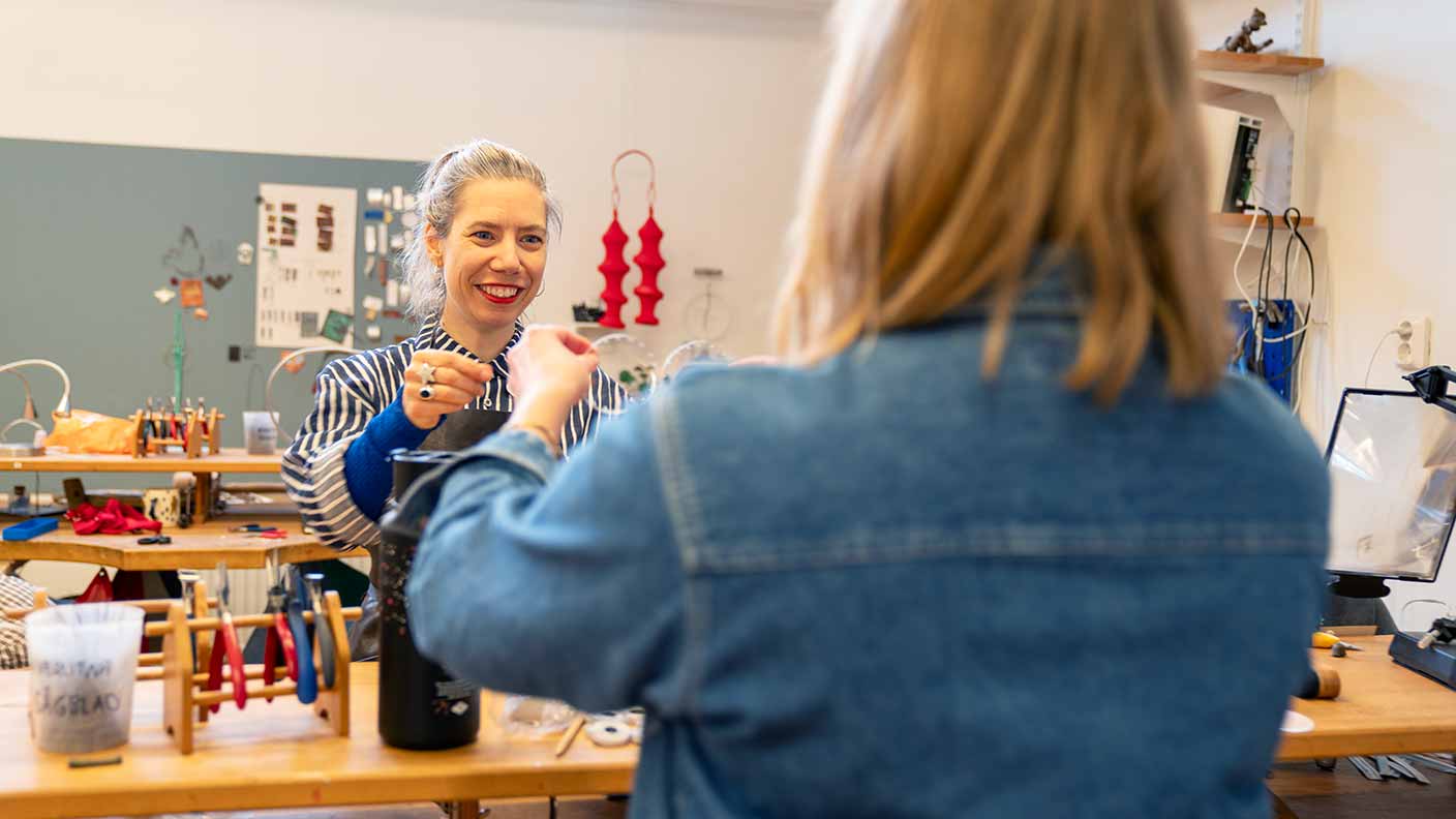 En person i silversmidesverkstaden tittat på något som en annan person, med ryggen mot kameran, visar upp. Runt om syns arbetsbänkar med verktyg och material. I bakgrunden hnger olika föremål i smyckekonst och smide på en ljusgrå tavla.