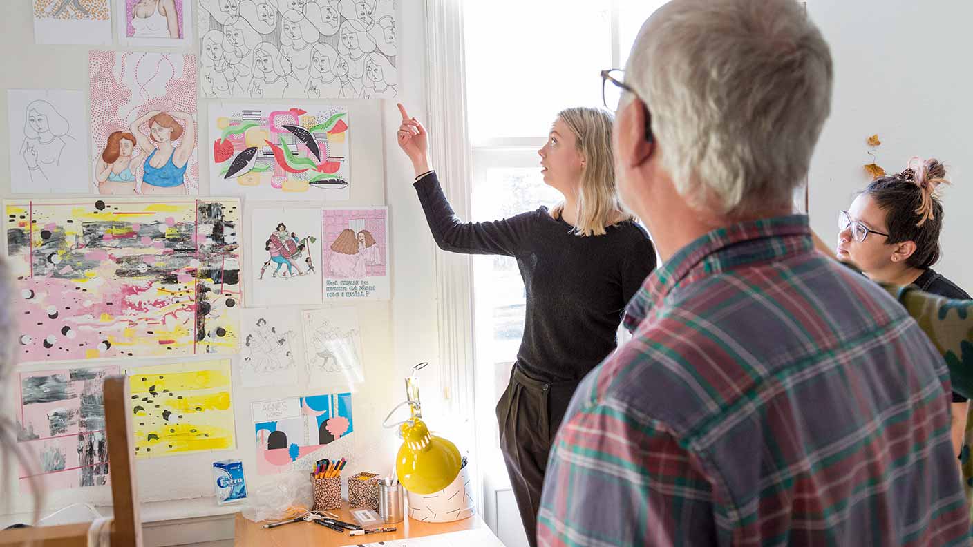 En kvinna med ljust hår och svarta kläder pekar på en serie teckningar och målningar som hänger på väggen. I förgrunden syns bakhuvudet på en person som lyssnar på hennes presentation.