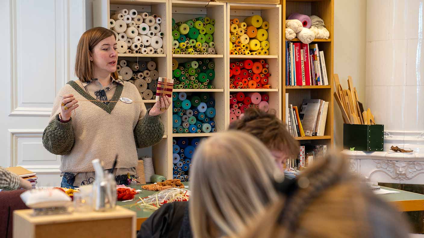 En lärare står framför en grupp deltagare och visar en vinda garn. I bakgrunden längs en vägg står tre stora hyllor fyllda med garnnystan i olika klara färger; grönt, gult, rött, blått med mera.