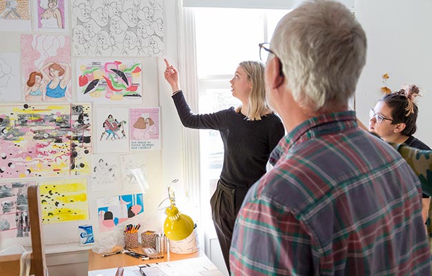 En kvinna med ljust hår och svarta kläder pekar på en serie teckningar och målningar som hänger på väggen. I förgrunden syns bakhuvudet på en person som lyssnar på hennes presentation.