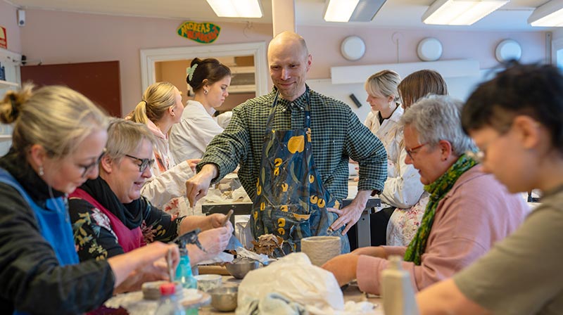 Andreas står vid ett bord i mitten av bilden. Han bär rutig skjorta och ett blått förkläde med gula figurer. Runt bordet sitter en grupp deltagare som fokuserar på att forma och modellera sina lerstycken.