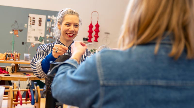 Anna är i silversmidesverkstaden; hon har på sig en randig skjorta och ett tjockt förkläde. Hon visar eller tittat på något som en deltagare framför henne i bild, med ryggen mot kameran, visar upp. Runt om syns arbetsbänkar med verktyg och material. I bakgrunden hnger olika föremål i smyckekonst och smide på en ljusgrå tavla.