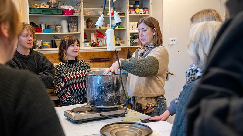 Lisa står vid ett bord omgiven av flera deltagare som ser på när hon visar en stor gryta på vad som verkar vara en portabel spis. Lisa är klädd i en stor stickad tröja som går i beige och grönt.