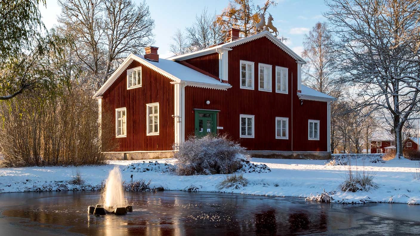 Skolmuseumet - ett rött trähus med vita knutar - i vinterskrud. På husets tak syns två lejon i trä. Framför huset, i bildens förgrund, ligger en damm med en aktiv fontän. 