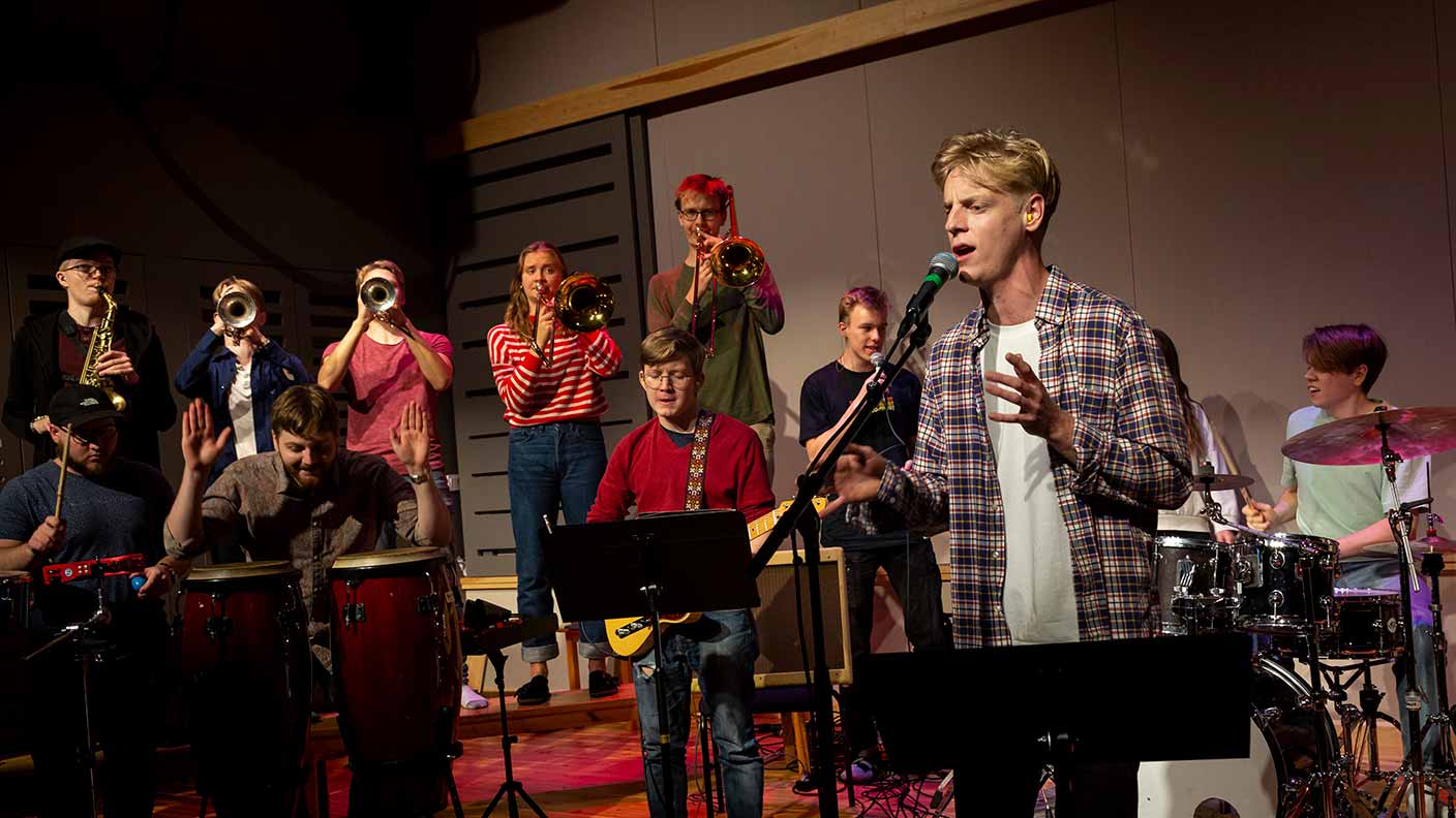 Ett stort band bestående av en blåssektion på fem personer, två percussionister, en gitarrist, en trummis, två bakgrundssångare och en leadsångare uppträder på en svagt upplyst scen.