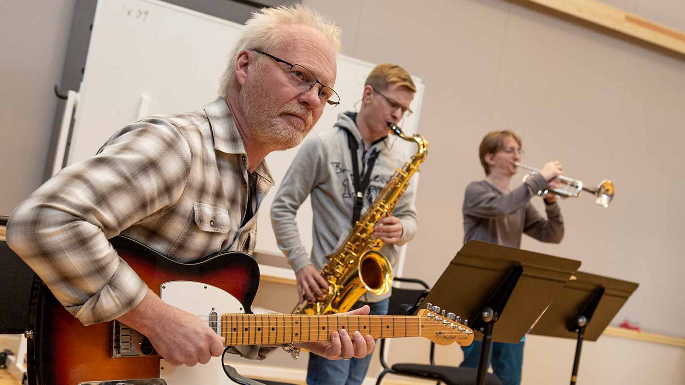 Tre personer musicerar i en stor och laturligt upplyst sal. I fögrunden sitter en gitarrist, som antas vara en lärare, och spelar samtidigt som han tittar på någon snett utanför bild. I bakgrunden syns två deltagare som spelar saxofon respektive trumpet.