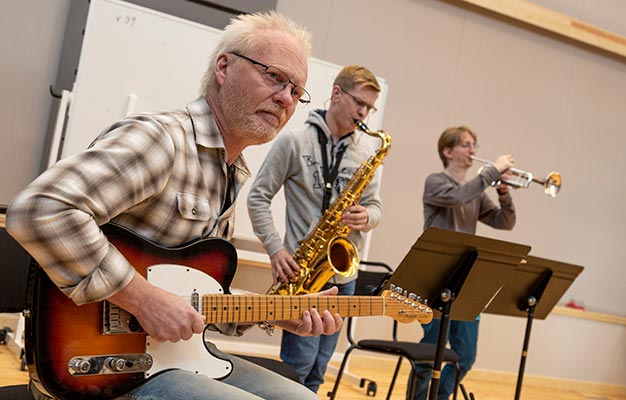 Tre personer musicerar i en stor och laturligt upplyst sal. I fögrunden sitter en gitarrist, som antas vara en lärare, och spelar samtidigt som han tittar på någon snett utanför bild. I bakgrunden syns två deltagare som spelar saxofon respektive trumpet.