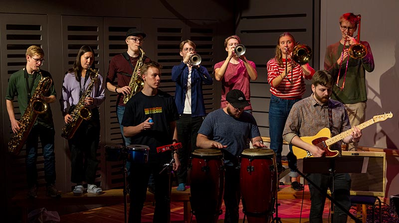 Tio musiker - en del av partybandet - uppträder på scen. I förgrunden står två personer som spelar percussion och en gitarrist. Bakom dem, på en upphöjning, står sju personer som spelar olika blåsinstrument; saxofon, trumpet och trombon.