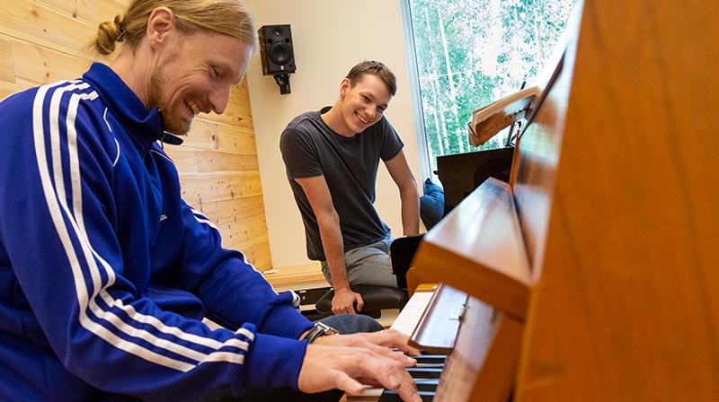 Vid ett piano i ljusbrunt trä sitter två personer; i förgrunden en lärare som demonstrerar något på piano och i bakgrunden en deltagare som tittar på. De är båda något böjda över pianot.