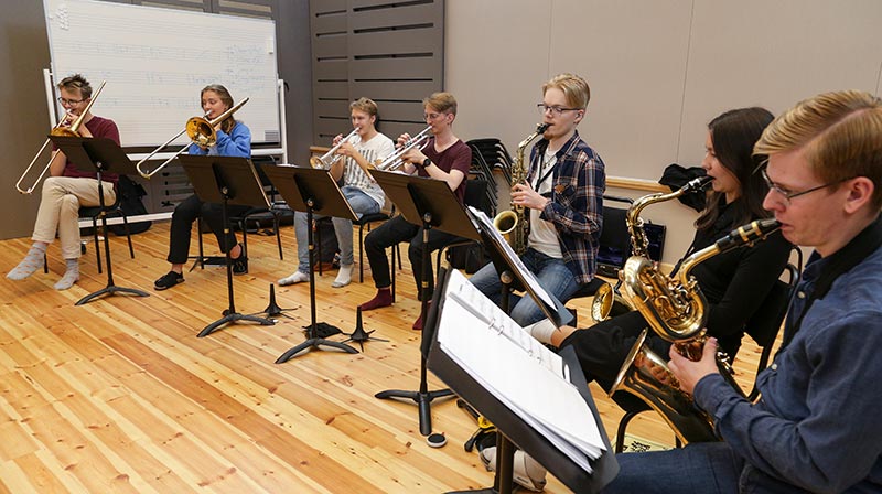 Sju personer sitter i en böjd rad på svarta stolar. De spelar olika blåsinstrument; trumpet, saxofon och trombon. De befinner sig i en stor och ljus lokal med högt i tak.